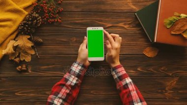 Sonbahar üstten görünüm kompozisyon. Defter, kitap, kazak, ahşap masa üzerinde akçaağaç yaprakları. Kadın kaydırma, yakınlaştırma beyaz smartphone ile yeşil ekran sayfalarında. Chroma anahtar