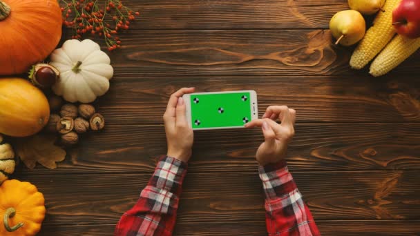 Φθινοπωρινά φρούτα και λαχανικά σύνθεση πάνω στο ξύλινο τραπέζι. Το Top view. Γυναίκα κύλιση, ζουμ, πατώντας στο λευκό smartphone με πράσινη οθόνη. Οριζόντια θέση. Κλειδί Chroma. Παρακολούθηση κίνησης — Αρχείο Βίντεο
