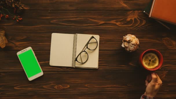 Autumn top view. Notobook, glasses, tea, white smartphone with green screen on the old wooden table. Woman drinking tea and taking glasses. Flat lay — Stock Video