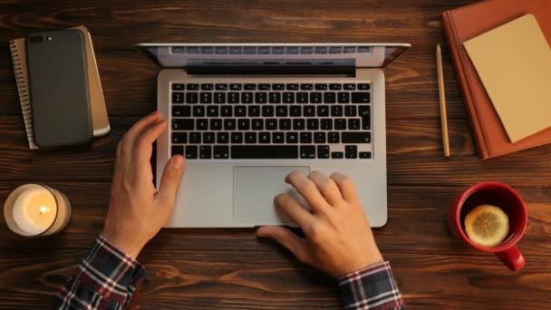 Vue de dessus de l'homme travaillant sur son ordinateur portable, tapant sur le clavier, surfant sur Internet. Fond de table en bois avec cahiers . — Video