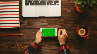 Renkli üstten görünüm. Kırmızı öğeler ahşap tablo yeşil ekranlı akıllı telefon kullanan kadın. Defter, çay bardağı, bitki ve laptop. Chroma anahtar. Hareket izleme