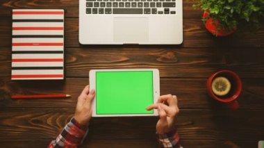 Kırmızı çay bardağı, renkli defter ve dizüstü bilgisayar ile ahşap tabloda yeşil ekranlı tablet aygıtı kullanan kadın. Chroma anahtar. Üstten Görünüm