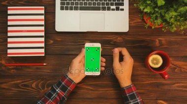 Hareket izleme ahşap tablo üzerinde yeşil ekran ile beyaz smartphone kullanan adam. Düz kırmızı çay Kupası, dizüstü bilgisayar ve dizüstü bilgisayar ile yatıyordu. Chroma anahtar