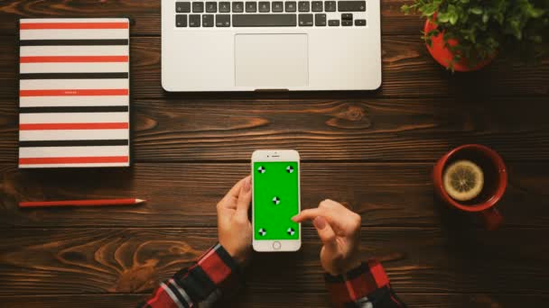 Vista superior. Mujer usando smartphone con pantalla verde en posición vertical en la mesa de madera con elementos rojos. Cuaderno, taza de té, planta y portátil. Clave de croma. Movimiento de seguimiento — Vídeo de stock