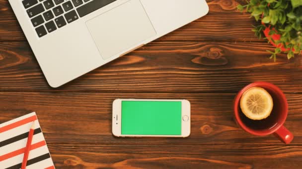 White smartphone with green screen in horizontal position on the wooden table with red items and laptop computer. Chroma key — Stock Video