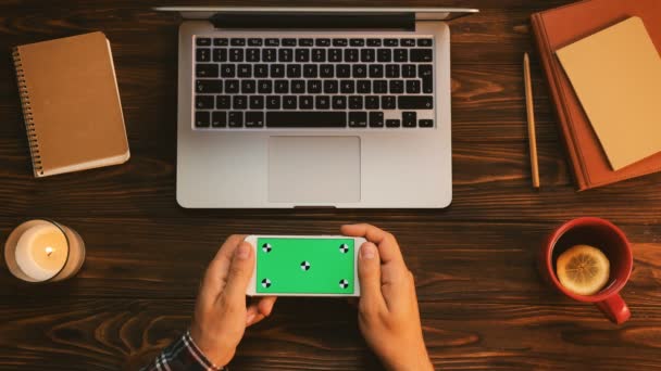 Hombre usando smartphone con pantalla verde en la mesa de madera con cuadernos, taza de té y vela. Vista superior. Clave de croma. Movimiento de seguimiento — Vídeos de Stock