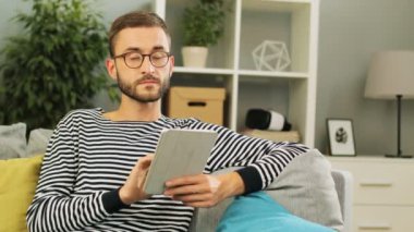 Dizüstü bilgisayar ve tninking evde mesajlara cevap hakkında yaşayan arka plan üzerinde kullanarak kanepede oturan gözlüklü yakışıklı genç beyaz adam. Kapalı atış.