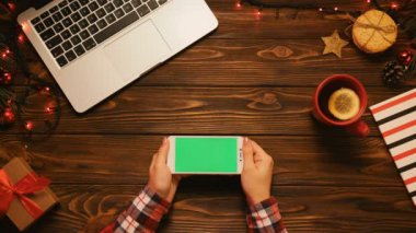 Ahşap masa üzerinde Noel üstten görünüm ile smartphone yeşil ekran, laptop, çay kupa ve Noel dekorasyonu ile. Kadın dokunarak, yakınlaştırma sayfaları. Chroma anahtar