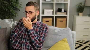 Gözlüklü genç adam hasta hissetme ve sürekli öksürük evde, kanepede oturuyor. Kapalı.