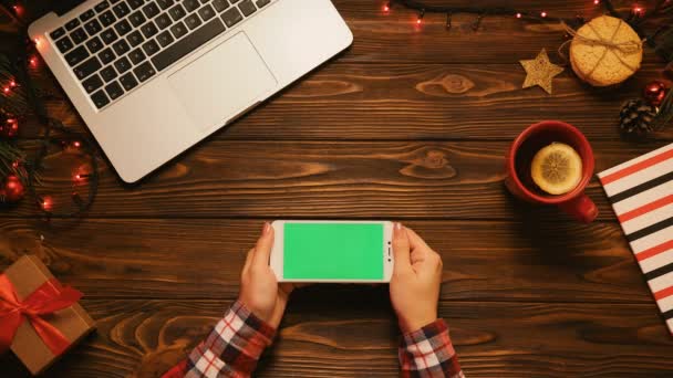 Ahşap masa üzerinde Noel üstten görünüm ile smartphone yeşil ekran, laptop, çay kupa ve Noel dekorasyonu ile. Kadın dokunarak, yakınlaştırma sayfaları. Chroma anahtar — Stok video