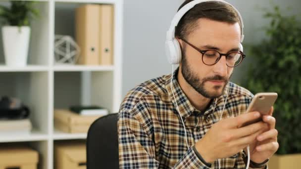 Close-up shot van een jonge Kaukasische zakenman met koptelefoon op, houden van mobiele telefoon in zijn handen, luisteren naar muziek in het moderne kantoor op een drukke dag. — Stockvideo