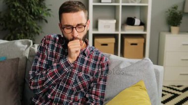 Gözlüklü genç çekici adam evde kanepede oturan, öksürme ve hapşırma. Kapalı atış.