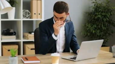 Rahatsızlık ve göz zorlanma duygu Ofis Masası kapalı gözlüklerini alarak ofiste, dizüstü bilgisayar üzerinde çalışan mutlu genç iş adamı. Kapalı atış. Yakın çekim.