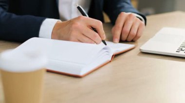 Beyaz sayfalarda yazmak için bir kalem kullanarak genç beyaz işadamı ile onun not defterinde yazılı bir modern ofis masasında oturan bir takım elbise eller yukarı kapatın.