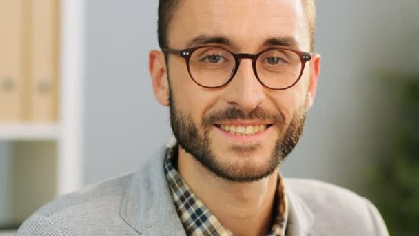 Nahaufnahme eines attraktiven kaukasischen Mannes mit Brille, der lässig in Hemd und grauem Hemd am Schreibtisch sitzt, in die Kamera blickt und lächelt.. — Stockvideo