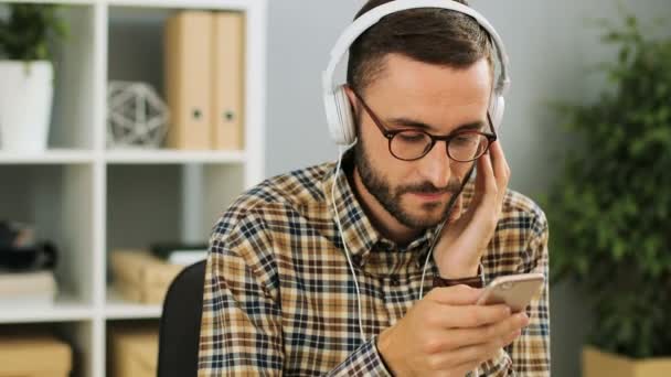 Κοντινό πλάνο του ένας χαρούμενος νεαρός επιχειρηματίας Καυκάσιος με ακουστικά, κρατώντας το τηλέφωνο στα χέρια του, να ακούτε μουσική στο σύγχρονο γραφείο σε μια πολυάσχολη ημέρα. — Αρχείο Βίντεο