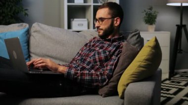 Sakallı genç beyaz adam kanepede yatan ve yaşam arka plan üzerinde bir dizüstü bilgisayarda akşam çalışma. Kapalı atış.