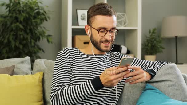 Atraktivní muž s sluchátka a mobil v rukou, usmívat a cítit uvolněně na gauči s polštářky na to na obývací pokoj pozadí. Vnitřní záběr. — Stock video