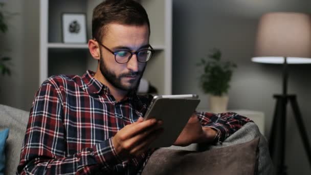Młody człowiek stylowy w cool sprawdzane koszula i modne okulary siedzi na kanapie w domu na tle życia i przy użyciu tabletu w czasie nocy. Wewnątrz strzał. — Wideo stockowe