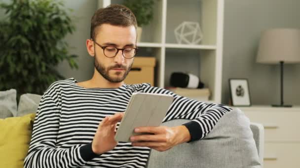 Красива біла людина в окулярах сидить на дивані, використовуючи планшет для роботи вдома в спокійній домашній атмосфері. Внутрішній постріл . — стокове відео