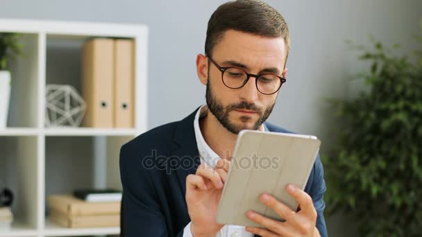 Retrato de um jovem homem de negócios caucasiano olhando para seu tablet ipad e trabalhando duro no escritório moderno. Indoor shot. Fechar . — Vídeo de Stock