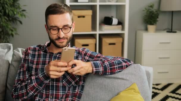 Jeune homme attrayant dans des lunettes est assis sur le canapé à la maison, tenant le téléphone portable et envoyer des SMS à sa petite amie via Internet, souriant et se sentant détendu. Intérieur . — Video