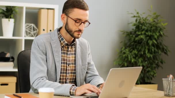 Atractivo hombre caucásico en gafas, con una camisa casual y una camisa gris sentado en el escritorio de la oficina, utilizando su computadora portátil para el trabajo. Disparo interior . — Vídeo de stock