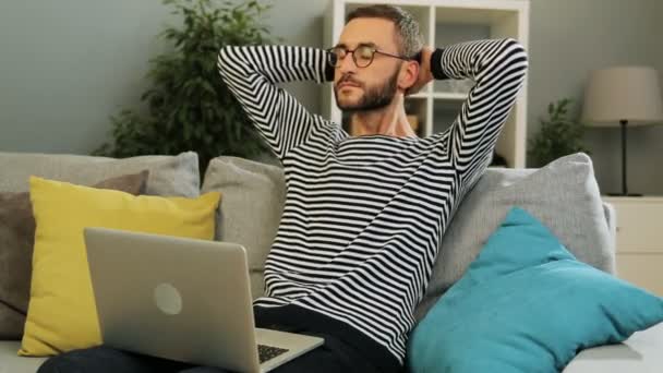 Stilig kaukasiska man i glasögon som sitter på soffan att skriva på den bärbara datorn och sedan puttinh hends över hans huvud att slappna av efter hårt arbete på hem bakgrunden. Inomhus skott. — Stockvideo