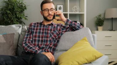 Gözlük ve rahat gömlek genç çekici adam cep telefonu tutan ve arkadaşlarıyla konuşurken evde, kanepede oturuyor. Kapalı.