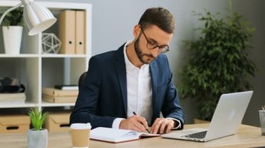 Çekici notlar trendy ofiste gözlüklü genç kendinden emin businessma. İşyerinde beyaz erkek oturma ve bir proje üzerinde çalışıyor.