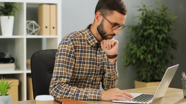 Jovem empresário concentrado sério em óculos que trabalham no escritório de arranque na moda. Homem caucasiano digitando no laptop sentado na mesa em um dia de trabalho ocupado . — Vídeo de Stock