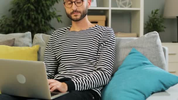 Exitoso hombre caucásico en gafas sentadas en el sofá utilizando el ordenador portátil para el trabajo, en casa sobre fondo vivo. Disparo interior . — Vídeos de Stock