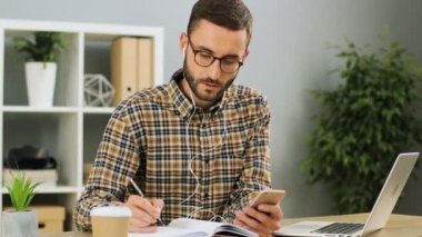 Gözlüklü, ciddi genç işadamı için Toplantı ve çalışma sert ofiste güneşli bir günde biranlaşma onun cep telefonundan notlar koymak için kulaklık kullanarak. Yakın çekim.