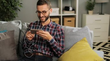 Checked rahat gömlek ve gözlük yakışıklı beyaz adam evde harika bir zamandı, kanepede oturan ve onun smartphone üzerinde kulaklık ile müzik dinlemek ve gülümseyerek. Kapalı atış.