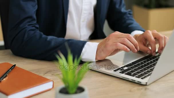 Close-up van de handen van een jonge Kaukasische zakenman in een pak zitten op het Bureau in een modern kantoor typen op zijn laptop, het maken van afspraken en het verzenden van e-mails. — Stockvideo