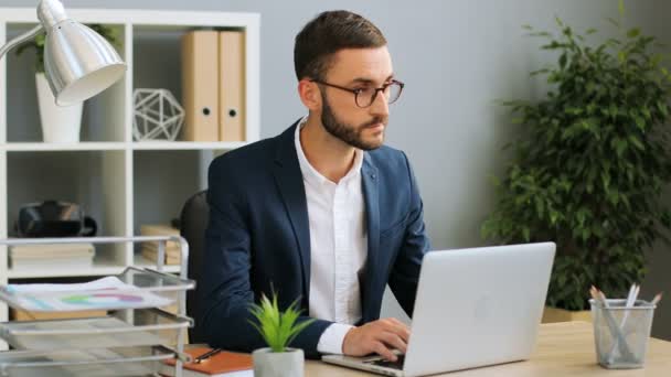 Egy jóképű üzletember szemüveg a munkahelyen, a trendi hivatalban ülő. Kaukázusi Férfi keresi a képernyő a laptop az asztalon. — Stock videók