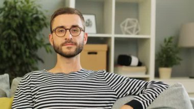 Bunu evde yaşayan arka plan üzerinde gülümseyen genç beyaz adam kanepede oturan ve kameraya, seyir bardaklarda. Kapalı atış.