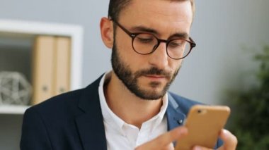 Onun smartphone office arka plan karşı yazarak genç şık işadamı gözlük, kadeh kapatın. Kapalı atış.