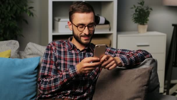 Close-up shot van een tevreden jonge man zittend op de bank terwijl het texting op de smartphone op de achtergrond van het leven in de avond. Binnen schot. — Stockvideo