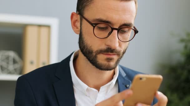 Nära upp skott av ung snygg affärsman i glasögon, att skriva på sin smartphone mot office bakgrund. Inomhus skott. — Stockvideo