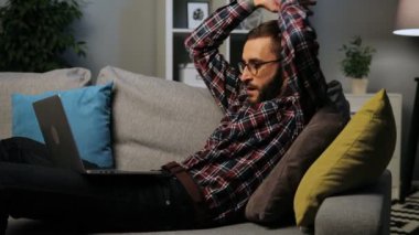 Sakallı genç beyaz adam kanepede yatan ve yaşam arka plan üzerinde bir dizüstü bilgisayarda akşam çalışma. Kapalı atış.