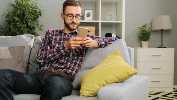 Aantrekkelijke jongeman in glazen zit op de Bank thuis, bedrijf van de mobiele telefoon en SMS zijn vrienden via internet. Indoor. — Stockvideo