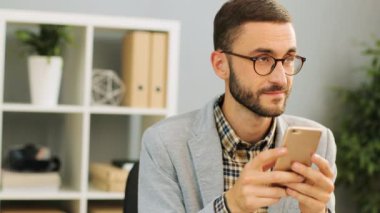 Fotoğraf üzerinde bir nessage düşünmek-e doğru onun smartphone tutarken yazmak sıradan bir gömlek ve ofis masa başında oturan gri gömlek giyen bir çekici beyaz adamın gözlükler, kapatın. Kapalı