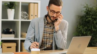 Telefonda konuşurken duygu mutlu ve memnun elinde kalem tutan neşeli bir adam ofis masa başında oturan gözlüklü atış kapatın. Kapalı atış.