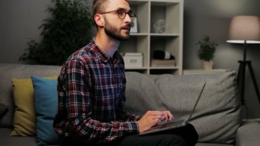 Evde meşgul ve düşünceli duygu genç beyaz adam bir dizüstü bilgisayarda çalışma kanepede oturan ekose desenli bir gömlek. Kapalı atış. Yakın çekim.