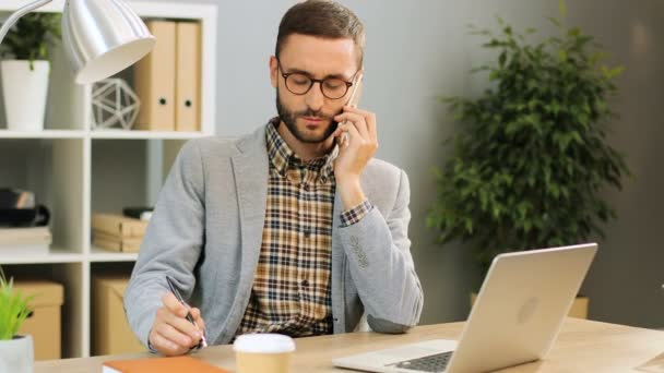 Nära upp skott av en glad man i glasögon som sitter vid det office-skrivbordet, prata i telefon och hålla en penna i handen. Inomhus skott. — Stockvideo