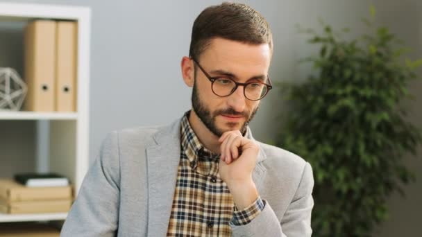 Foto ravvicinata di un uomo allegro con gli occhiali seduto alla scrivania dell'ufficio, che guarda in basso e sorride alla telecamera. Indoor shot . — Video Stock