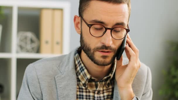 Primer plano de un hombre feliz con gafas sentado en el escritorio de la oficina, hablando por teléfono y sintiéndose contento y satisfecho durante la conversación con un colega. Disparo interior . — Vídeos de Stock