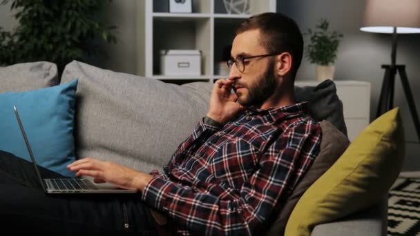 Kaukasische jongeman liggend op de Bank bezig met een laptop en praten over de smarthone, gezellige thuis voelen. Binnen schot. Close-up. — Stockvideo