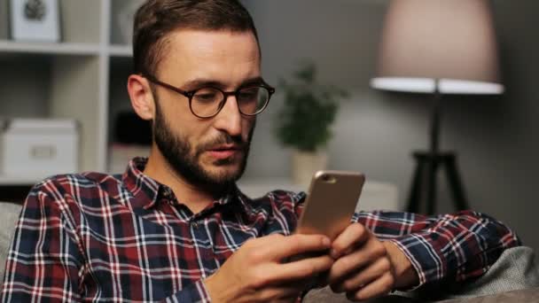 Primer plano de un joven feliz con gafas sentado en el sofá mientras envía mensajes de texto en el teléfono inteligente en el fondo borroso por la noche. Disparo interior . — Vídeos de Stock